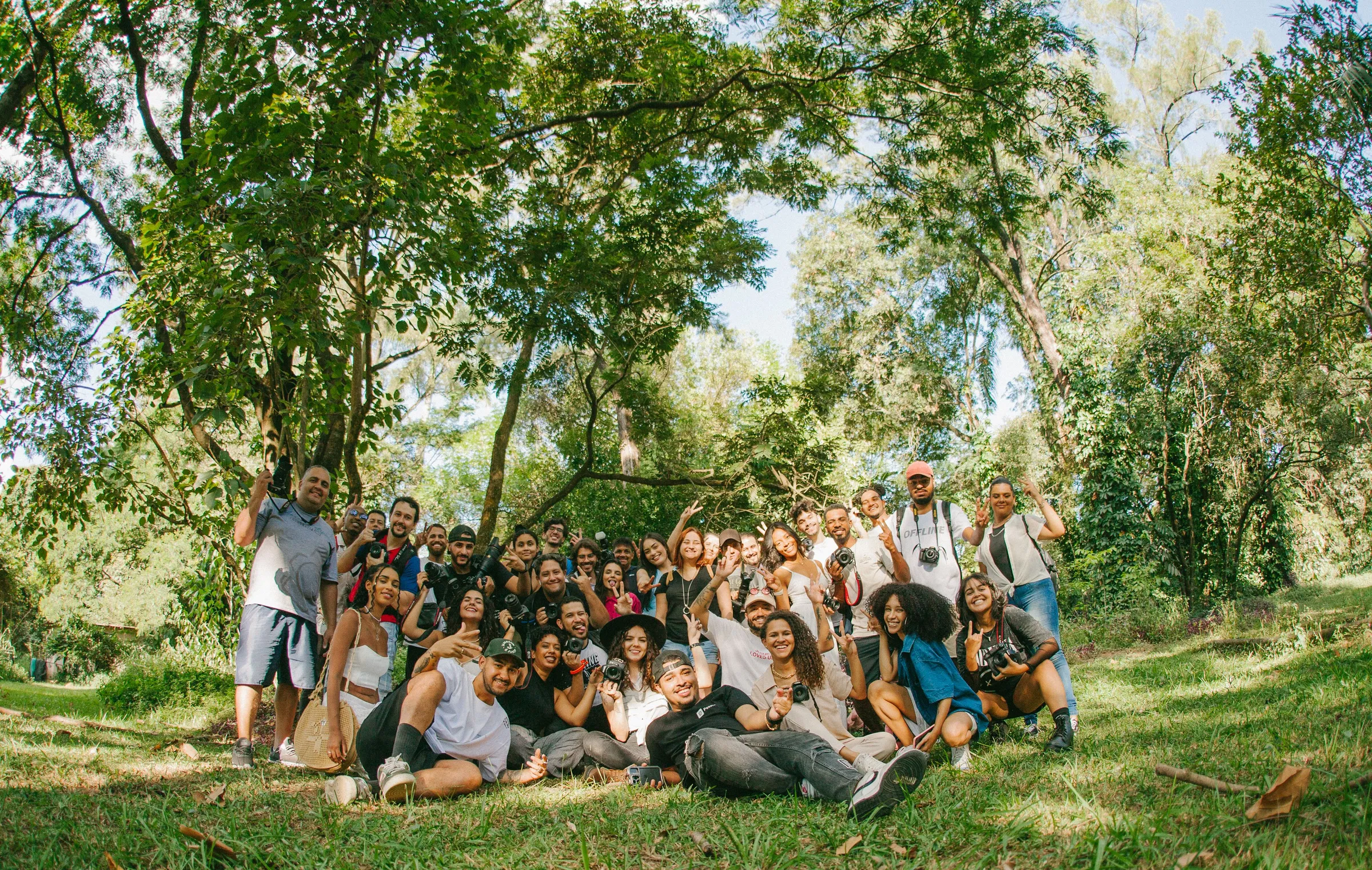 Un grupo numeroso de personas sonríe y saluda a la cámara reunido bajo los árboles de un parque